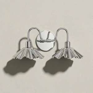Front view of a polished chrome double wall sconce with symmetrical curved arms, a round backplate, and two downward-facing scalloped shades casting distinct shadows.