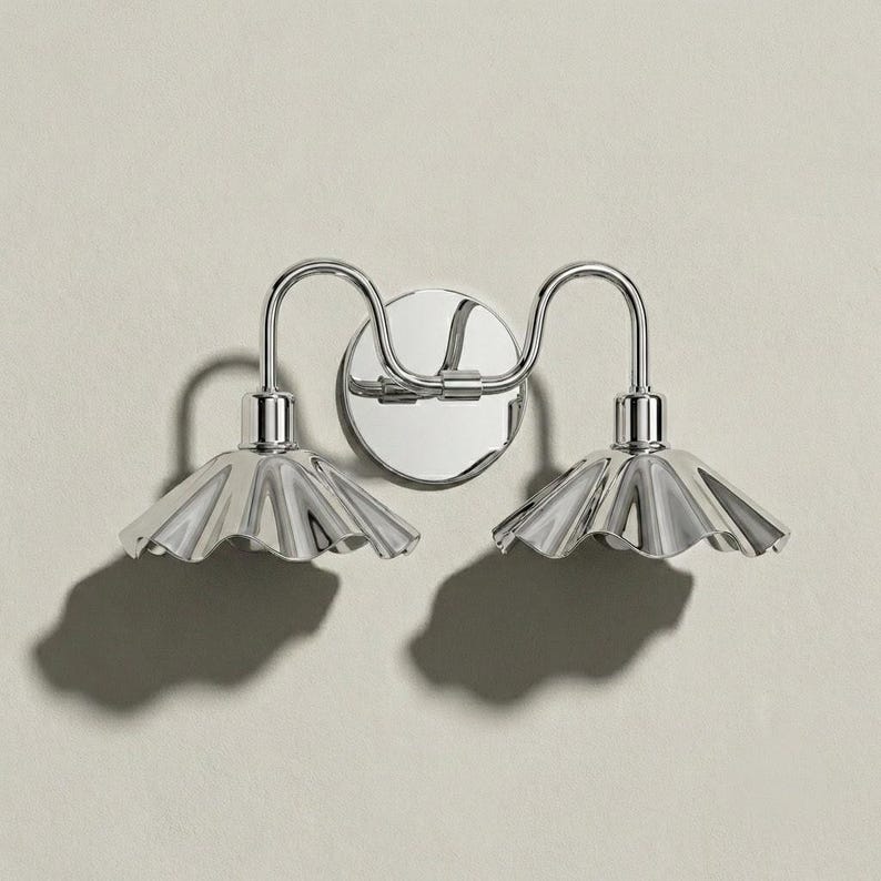 Front view of a polished chrome double wall sconce with symmetrical curved arms, a round backplate, and two downward-facing scalloped shades casting distinct shadows.