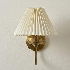May include: A wall-mounted sconce with a pleated, off-white lampshade. The fixture has a brass finish, including the circular backplate, the curved arm, and the decorative details. The lampshade is cone-shaped and made of a textured material.