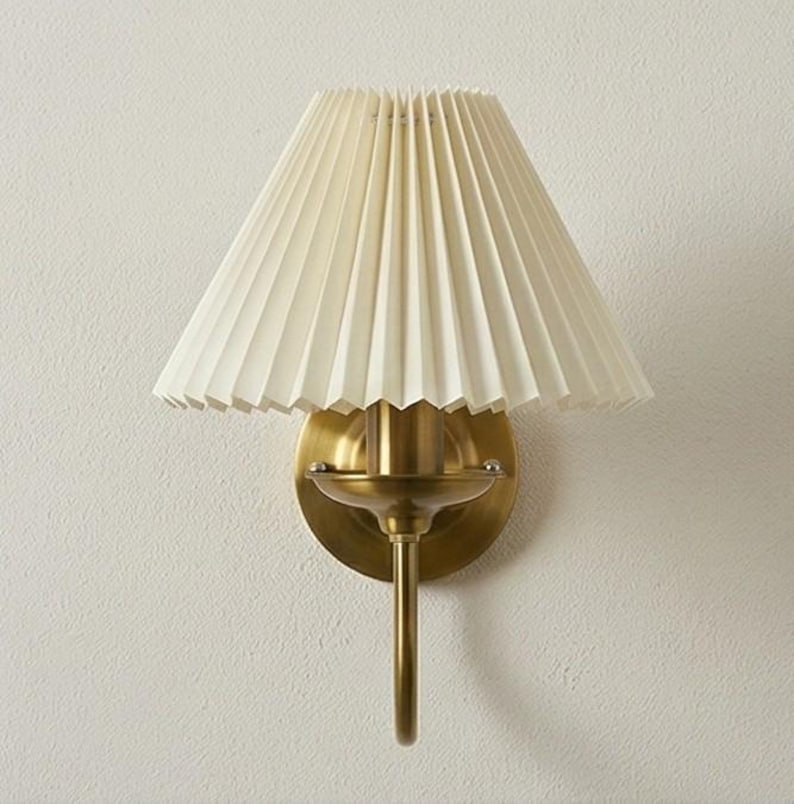 May include: A wall-mounted sconce with a pleated, off-white lampshade. The fixture has a brass finish, including the circular backplate, the curved arm, and the decorative details. The lampshade is cone-shaped and made of a textured material.