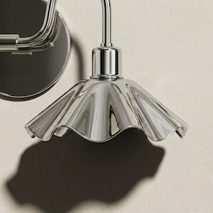 Close-up of a polished chrome wall sconce shade showing its reflective metal surface, radiating raised flutes, scalloped rim, cylindrical socket holder, and curved arm.