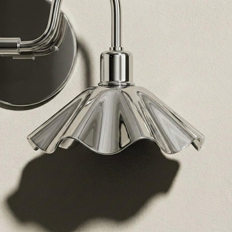 Close-up of a polished chrome wall sconce shade showing its reflective metal surface, radiating raised flutes, scalloped rim, cylindrical socket holder, and curved arm.