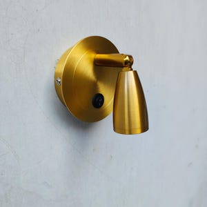 Puede incluir: Un foco de pared en oro cepillado que cuenta con una placa base circular con un pequeño interruptor negro y un cabezal de lámpara cónico ajustable unido por un brazo horizontal corto, proporcionando una fuente de luz direccional.