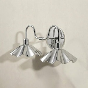Angled side view of a double-arm chrome wall light showing its curved tubular arms, circular mounting plate, reflective finish, and sculpted fluted shades.