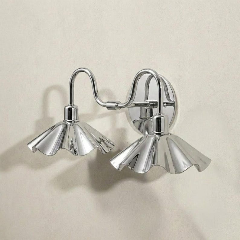 Angled side view of a double-arm chrome wall light showing its curved tubular arms, circular mounting plate, reflective finish, and sculpted fluted shades.