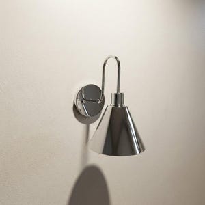 Puede incluir: Aplique de pared cromado pulido con una pantalla cónica. La lámpara tiene un brazo curvo que conecta la pantalla a un soporte de pared circular. El aplique está montado en una pared de color beige claro, proyectando una sombra.