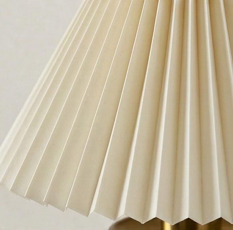 May include: Close-up of a cream-colored pleated lampshade. The shade has a classic design with vertical folds, creating a textured surface. The color is a soft, warm off-white, suitable for various interior styles.