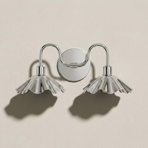 Pode incluir: Arandela de parede cromada polida com duas luzes. Cada luz tem uma sombra com babados em forma de pétala. A luminária é montada em uma placa traseira circular e projeta uma sombra na parede de cor neutra.