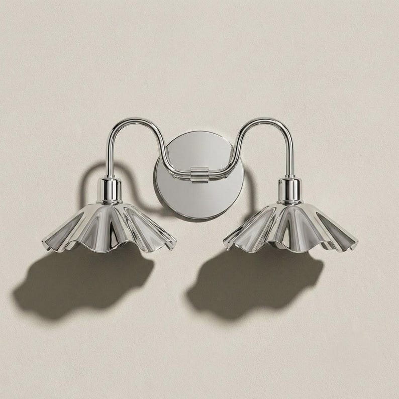 Double wall sconce with polished chrome curved arms, a round wall plate, and two reflective fluted shades with scalloped edges, mounted on a lightly textured off-white wall.