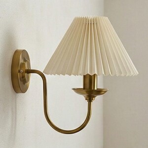 May include: A wall-mounted sconce with a pleated cream-colored lampshade. The fixture has a brass finish, with a curved arm and a circular base. The design is elegant and suitable for interior lighting.