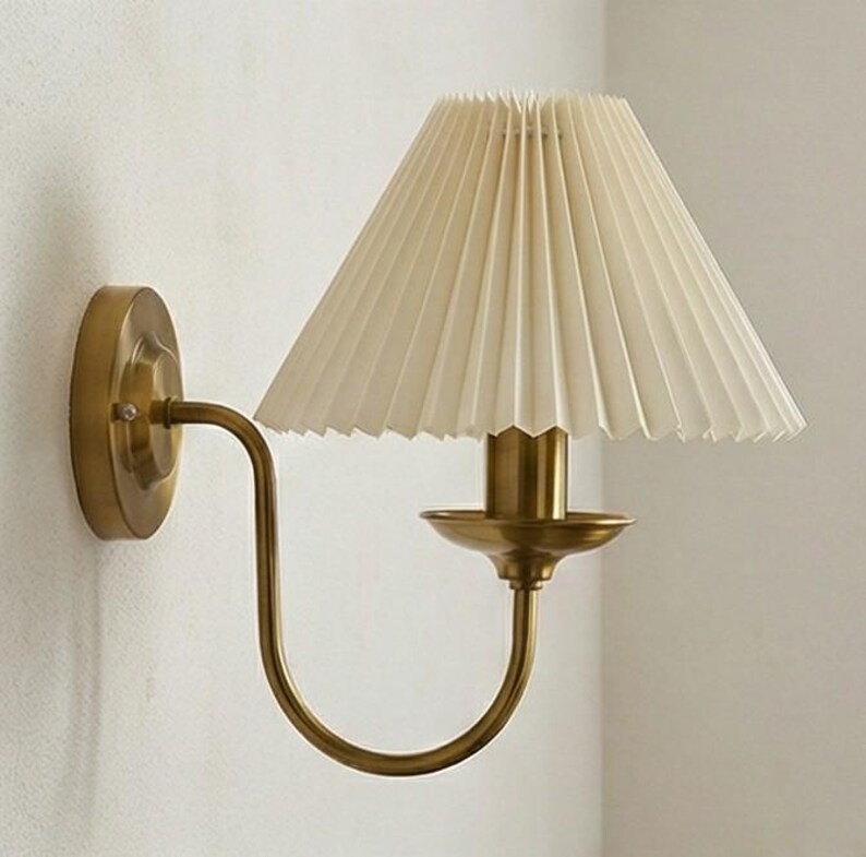 May include: A wall-mounted sconce with a pleated cream-colored lampshade. The fixture has a brass finish, with a curved arm and a circular base. The design is elegant and suitable for interior lighting.