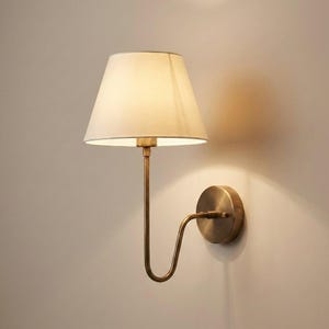 Puede incluir: Una lámpara de pared con una pantalla de color crema y un marco de metal color latón. La lámpara tiene un brazo curvo y una base circular. La pantalla es cónica y dirige la luz hacia abajo. Diseñada para uso en interiores.