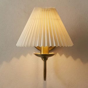May include: A wall-mounted lamp with a pleated white lampshade and a brass-colored base. The lamp has a long, slender stem and a small, circular dish beneath the shade. The light casts a shadow on the wall.