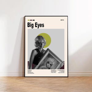 May include: A framed movie poster for "Big Eyes" (2014) featuring a woman holding a painting of a child with large eyes. The poster has a cream background with the title in bold black letters and a green circle.