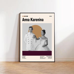 May include: A framed movie poster for "Anna Karenina" (2012) featuring a black and white image of two people holding hands. The poster has a beige background with a brown and burgundy accent. The title is in bold black text.