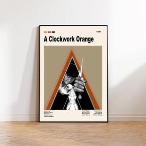 Op de afbeelding: Een ingelijste poster voor de film "A Clockwork Orange" (1971). De poster toont een zwart-wit afbeelding van een man die een wapen vasthoudt, tegen een bruine en oranje driehoek. De filmtitel staat in vette zwarte letters.
