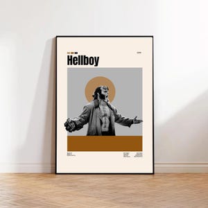 May include: A framed poster featuring the word "Hellboy" in black text. The poster has a beige background with a gray rectangle and a brown horizontal bar. The image depicts a black and white illustration of Hellboy with his arms outstretched.