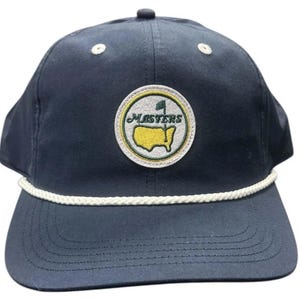 Può includere: Un cappellino da baseball blu navy con una toppa circolare ricamata sul davanti. La toppa mostra una mappa gialla degli Stati Uniti con una bandiera verde e la scritta "MASTERS" in verde. Un cordoncino bianco intrecciato decora la visiera.