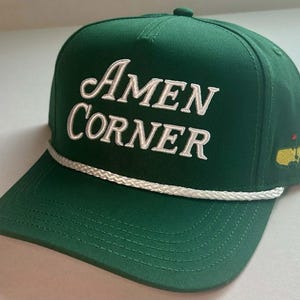 Può includere: Un cappellino da baseball verde con la scritta "Amen Corner" ricamata in bianco. Il cappellino presenta un dettaglio in corda bianca lungo la tesa e un piccolo logo sul lato.