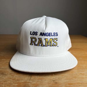 May include: A white baseball cap featuring the embroidered text LOS ANGELES in dark blue and RAMS in yellow with a dark blue outline. The cap has a structured crown, a flat brim with visible stitching, and a small trademark symbol at the end.