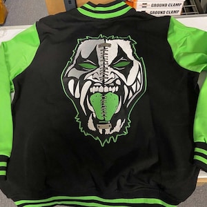 Könnte beinhalten: Schwarz-grüne Collegejacke mit einem großen Monstergesicht-Motiv auf dem Rücken. Die Ärmel sind leuchtend grün, und der Kragen, die Manschetten und der Saum weisen grün-schwarze Streifen auf. Das Monstergesicht hat grüne Augen und Zunge.