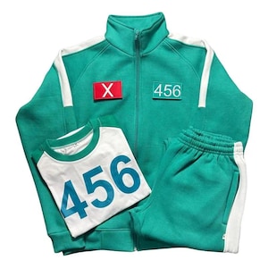 Op de afbeelding: Een tealkleurige trainingspakset met een jas met ritssluiting en witte strepen, een bijpassende joggingbroek en een wit T-shirt. Op de jas en het T-shirt staat het nummer "456" gedrukt en de jas heeft een rode patch met de letter "X".