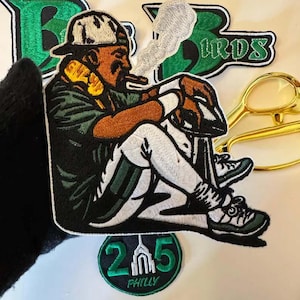 May include: An embroidered patch featuring a figure wearing a cap, dark shirt, and white trousers, smoking a cigar with visible smoke. The patch includes a smaller circular patch with the text 215 PHILLY and a larger patch with the text BIRDS.