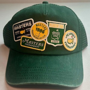 Può includere: Un cappellino trucker verde scuro con cinque toppe ricamate relative al Masters Tournament. Le toppe includono testi come Masters 1986, Masters Tournament e Augusta National Golf Club, insieme a varie grafiche a tema golfistico.