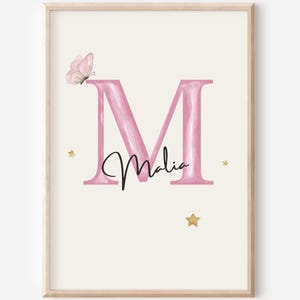 Könnte beinhalten: Ein gerahmter Kunstdruck mit einem großen, aquarellartigen rosa Buchstaben M auf cremefarbenem Hintergrund. Der Name Malia ist in schwarzer Schreibschrift über den Buchstaben geschrieben. Ein kleiner rosa Schmetterling und drei goldene Sterne.