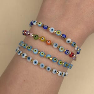 Puede incluir: Cuatro pulseras de cuentas con un diseño de ojo turco. Las pulseras presentan pequeñas cuentas redondas de varios colores, incluyendo azul, blanco, amarillo y rojo, ensartadas en una cadena dorada. Las pulseras se llevan en una muñeca.