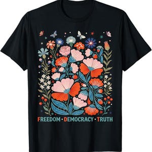 Op de afbeelding: Zwart t-shirt met een bloemenontwerp met roze, oranje en blauwe bloemen, groene bladeren en vlinders. De woorden "FREEDOM • DEMOCRACY • TRUTH" staan onderaan het ontwerp.