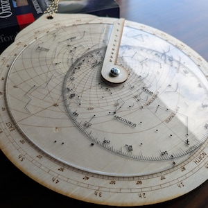 Könnte beinhalten: Ein hölzernes Astrolabium mit eingravierten Sternkarten und Gradmarkierungen. Das Instrument hat eine rotierende zentrale Scheibe und eine transparente, kreisförmige Auflage. Das Wort "ARCUTURUS" ist sichtbar. Ein Buch ist teilweise im Hintergrund zu sehen.
