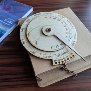 Könnte beinhalten: Ein hölzernes Astrolabium mit Zeiger, graviert mit Monaten, Daten und Sternbildern. Das Gerät befindet sich auf einer braunen Pappschachtel, im Hintergrund ein Buch mit dem Titel "STARS".