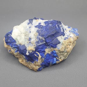 May include: A raw, natural mineral specimen featuring a striking combination of deep blue lapis lazuli, white quartz, and golden pyrite. The stone has a rough, irregular shape and a mix of textures.