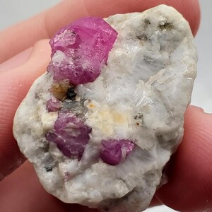 May include: A raw, uncut ruby gemstone cluster. The ruby crystals are a vibrant pinkish-red color, set against a matrix of white and gray rock. The rough texture of the stone is visible, showcasing its natural form.