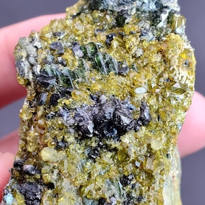 May include: Close-up of a mineral specimen featuring a mix of colors and textures. The rock displays a combination of yellow-green, black, and dark green crystals. The surface is rough and uneven, with a variety of crystal formations.