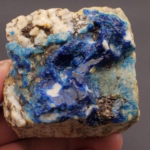 May include: A close-up of a raw mineral specimen featuring a striking contrast of colors and textures. The stone displays a deep, vibrant blue mineral formation against a backdrop of beige and white rock. Small flecks of metallic minerals add sparkle.