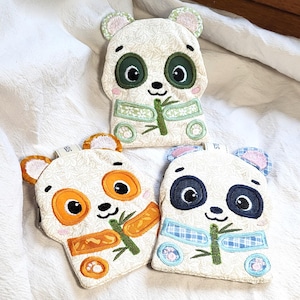 Peut inclure: Trois pochettes en tissu en forme de panda avec des détails brodés. Chaque panda a une palette de couleurs différente : vert, orange et bleu. Ils présentent des tiges de bambou et des motifs floraux, avec un fond crème et un tissu texturé.