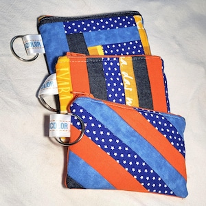 Peut inclure: Trois pochettes zippées colorées avec des porte-clés. Les pochettes présentent un motif patchwork avec des rayures bleues, orange et jaunes, et certaines ont des motifs d'étoiles blanches. Chaque pochette a un ruban blanc avec le mot "COLOR" imprimé dessus.