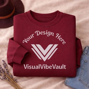 May include: A maroon crewneck sweatshirt with the text "Your Design Here" and "VisualVibeVault" in white. A white V-shaped logo is centered below the text. The sweatshirt is folded, with a brown hat and blue jeans visible in the background.
