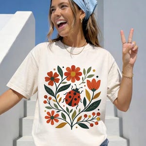 May include: Cream-colored t-shirt featuring a floral and ladybug design. The graphic includes red and orange flowers, green leaves, and a red ladybug with black spots. The shirt has short sleeves and a relaxed fit.