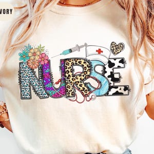 May include: Ivory-colored sweatshirt with the word "NURSE" in a colorful, patterned design. The "N" is leopard print with floral accents, the "U" is purple, the "R" is leopard print, the "S" is blue, and the "E" is cow print. A syringe, nurse's hat, and heart are also featured.
