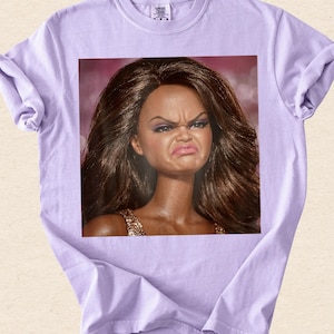 May include: A light purple short-sleeved t-shirt featuring a square graphic print on the chest. The graphic depicts a doll with long, dark brown hair and a facial expression showing a scrunched nose and pursed lips, wearing a gold-toned top.