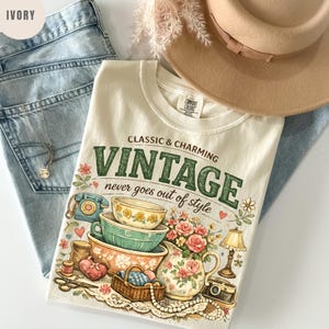 May include: An ivory t-shirt featuring a graphic print with the text CLASSIC & CHARMING VINTAGE never goes out of style. The illustration includes stacked bowls, a floral pitcher, a rotary phone, a lamp, a camera, and sewing supplies.