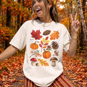 May include: A cream-colored short-sleeve t-shirt featuring a central graphic print of various autumn elements including a red maple leaf, orange pumpkins, pinecones, acorns, a red-capped mushroom, a brown mushroom, and yellow oak leaves.