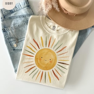 May include: An ivory t-shirt featuring a watercolor-style yellow sun graphic with a smiling face and multi-colored rays in shades of orange, brown, and sage green. The shirt is paired with light blue denim jeans, a tan wide-brim hat, and a silver necklace.