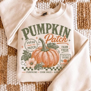 May include: A cream-colored crewneck sweatshirt featuring a graphic print with the text PUMPKIN Patch, FRESH FROM THE PATCH, FARM FRESH, LOCALLY GROWN, AUTUMN, PUMPKINS, CORN MAZE, and APPLE. The design includes a large orange pumpkin and leaves.