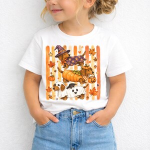 May include: A white short-sleeve t-shirt featuring a graphic of three dachshunds in Halloween costumes. One dog wears a purple star-patterned witch hat and cape, one is dressed as a pumpkin, and one wears a ghost sheet with bat wings.