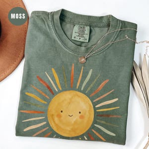 May include: A moss green short-sleeve t-shirt featuring a watercolor-style sun graphic with a smiling face. The sun has rays in shades of yellow, orange, and muted green. The shirt has a Comfort Colors label at the neck and a thin gold necklace.