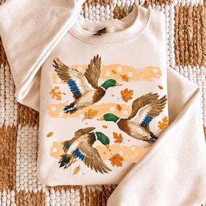 May include: A cream-colored crewneck sweatshirt featuring a graphic print of three mallard ducks in flight. The ducks have green heads, brown bodies, and blue wing patches. The design includes scattered orange autumn leaves and light orange stripes.